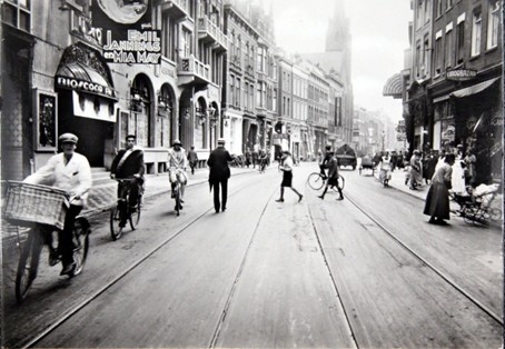Drukke Rotterdamse straat in het begin van de 20e eeuw met fietsers, voetgangers en winkelpuien, nabij het oude Luxor.