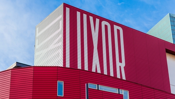 De gevel van het Luxor-gebouw is zichtbaar met een rode bekleding en grote witte verticale letters die samen het woord “LUXOR” vormen.