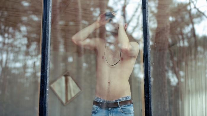 Man in spijkerbroek en ontbloot bovenlijf staat voor het raam en kijkt door een verrekijker.