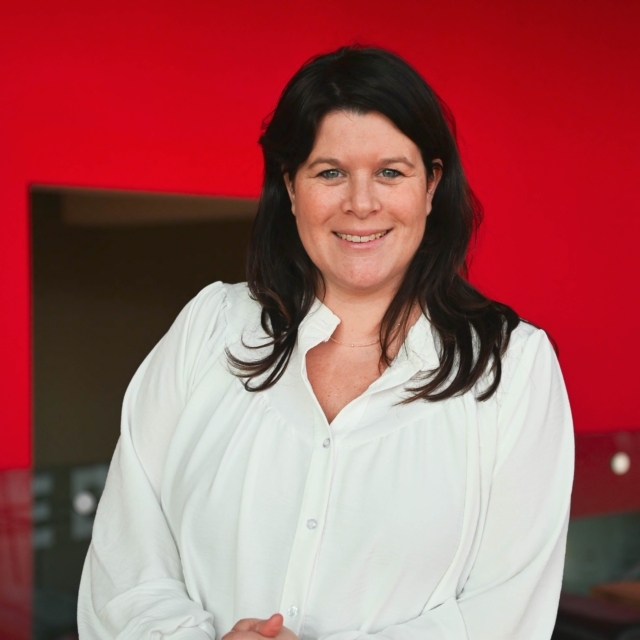 Vrouw met zwart lang haar en een witte blouse kijkt de camera in.