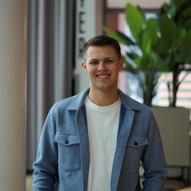 Man met wit t-shirt en een spijkerblouse kijkt lachend de camera in.