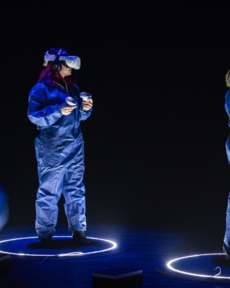 Twee personen in blauwe overalls staan in verlichte cirkels in een donkere ruimte terwijl ze VR-headsets en controllers dragen, verdiept in een virtuele ervaring.