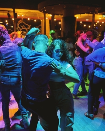 Een groep mensen danst in paren op een drukke dansvloer; in het midden danst een persoon in een kort wit shirt met een partner in een blauw shirt, terwijl verschillende andere dansende koppels hen omringen.