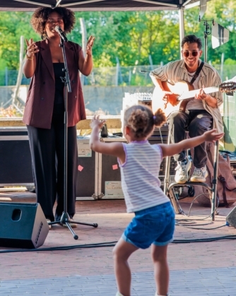 Een vrouw zingt enthousiast in een microfoon terwijl een man naast haar gitaar speelt, en een jong kind voor het podium vrolijk danst