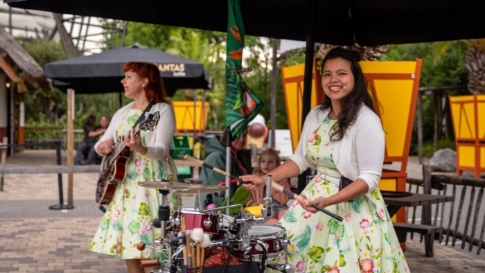 Twee vrouwen in zomerse jurken met bloemenpatroon spelen muziek in een buitenlocatie; de een bespeelt een gitaar en de ander een klein drumstel terwijl ze naar de camera lacht, met parasols en bezoekers op de achtergrond.