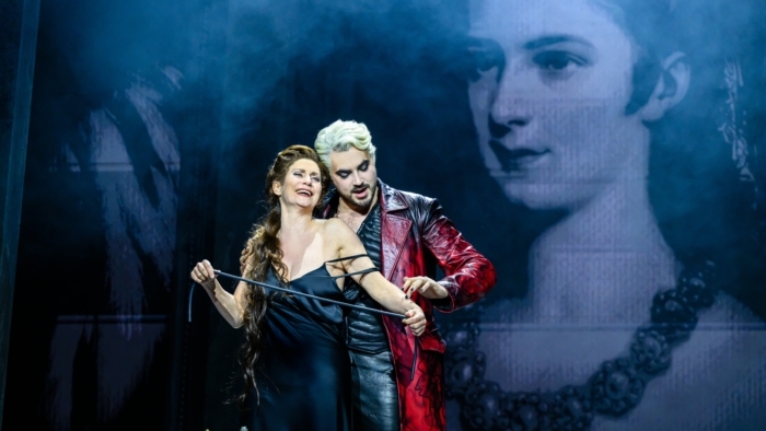 Een vrouw in een zwarte jurk staat vooraan terwijl een man in een lange rode jas achter haar staat en haar armen vasthoudt, beiden midden in een expressieve toneelscène met een groot zwart-wit portret van een vrouw op de achtergrond.