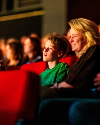 Een jongen in een groene trui en een vrouw zitten naast elkaar in rode theaterstoelen en kijken aandachtig voor zich uit, met andere bezoekers om hen heen.