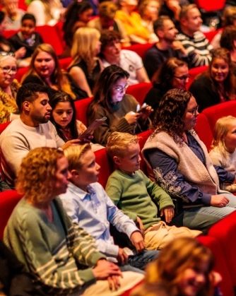 Een grote groep volwassenen en kinderen zit in rode theaterstoelen, waarbij verschillende bezoekers met elkaar praten, een programmablad bekijken of op hun telefoon kijken.